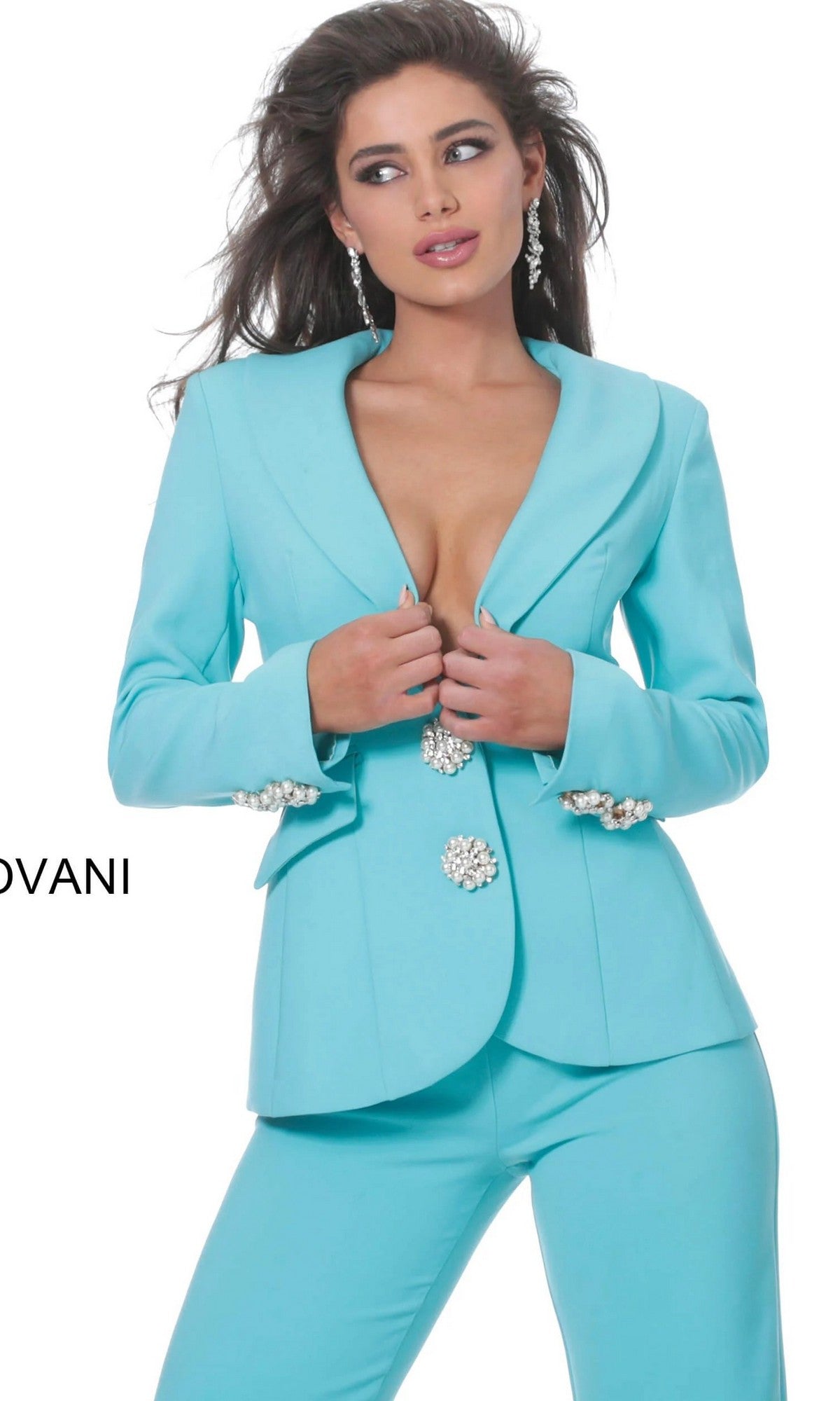 A woman wearing the Jovani 02637 Long Jumpsuit—a fitted light blue jumpsuit with jeweled buttons and earrings—stands against a white background.